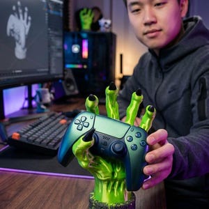 May include: A teal and black video game controller held by a green monster hand-shaped stand. The stand has long green fingers with black fingernails. The controller has buttons and a touchpad.