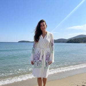 Peut inclure: Robe blanche avec broderie de lavande violette sur les manches et l'ourlet. La robe a une ceinture assortie et un ourlet à franges. La personne se tient sur une plage de sable avec l'océan en arrière-plan.