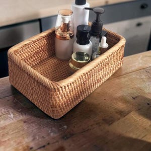 May include: A rectangular woven rattan basket with rounded corners containing several skincare bottles and jars. The basket features a light brown natural finish with a tight, uniform weave pattern and a thick, braided rim along the top edge.