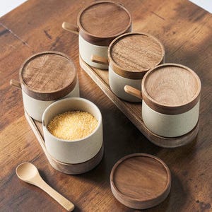 May include: A set of six ceramic spice jars with wooden lids and handles, arranged on a wooden tray. One jar is open and filled with yellow granules. A small wooden spoon and a spare lid are also present on a wooden surface.