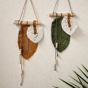 May include: Two macrame feather wall hangings. One is brown with a white accent, the other is green with a white accent. Each has a wooden dowel and hanging cord. Decorative wooden beads are attached to the bottom.