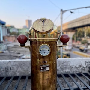 Pode incluir: Uma bússola magnética de latão vintage com um topo redondo e um corpo cilíndrico. Possui um pequeno medidor e duas esferas vermelhas em cada lado. A bússola tem uma aparência desgastada, sugerindo idade e uso. O texto "MAGNETIC COMPASS" é visível.