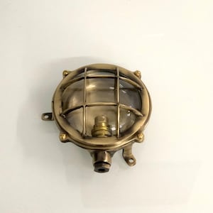 May include: A round, antique brass-colored wall-mounted light fixture. The fixture has a clear, domed glass cover protected by a grid. It features a bulb socket and mounting brackets, suggesting a nautical or industrial design.