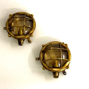 Puede incluir: Dos apliques de pared redondos de color latón. Cada lámpara tiene una cubierta de vidrio transparente y redondeada protegida por una rejilla. Las lámparas tienen una estética vintage y náutica, con una base pequeña y cilíndrica y soportes de montaje.