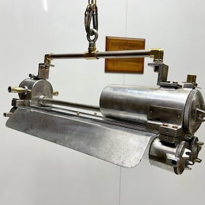 Könnte beinhalten: Industrielle Metallleuchte mit gebürsteter Oberfläche. Die Leuchte hat einen rechteckigen Schirm und zylindrische Komponenten, die durch Messingfittings und Metallrohre verbunden sind. Sie wird an einer Kette und einem Karabinerhaken aufgehängt.