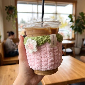 Puede incluir: Un portavasos de ganchillo rosa y verde con una flor blanca y rosa. El portavasos está envuelto alrededor de una taza de plástico transparente llena de café helado. La etiqueta en el portavasos dice "pixelclover".