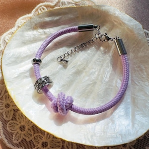May include: A lavender rope bracelet with silver-tone metal accents. The bracelet features a knot design and a decorative ring with clear rhinestones. The bracelet is displayed on a white, shell-shaped surface.