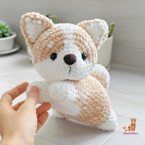 May include: A plush crochet corgi toy made of soft, fuzzy yarn in shades of cream and light tan. The stuffed animal features pointed ears, a dark brown stitched nose, and black plastic eyes. A small logo with the text ChenPattern is visible.