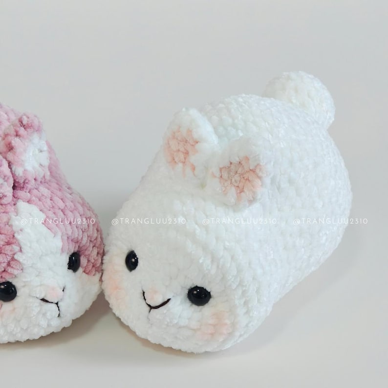 May include: Two plush crochet stuffed animals shaped like round bunnies. One bunny is white with light pink inner ears and cheek accents, while the other is dusty pink and white. Both feature black plastic eyes and embroidered noses.