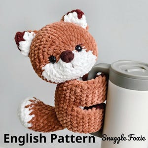 May include: A crocheted fox plushie in shades of brown, white, and burgundy, holding a white and gray travel mug. The fox has black eyes and a brown nose. The text "English Pattern Snuggle Foxie" is at the bottom.