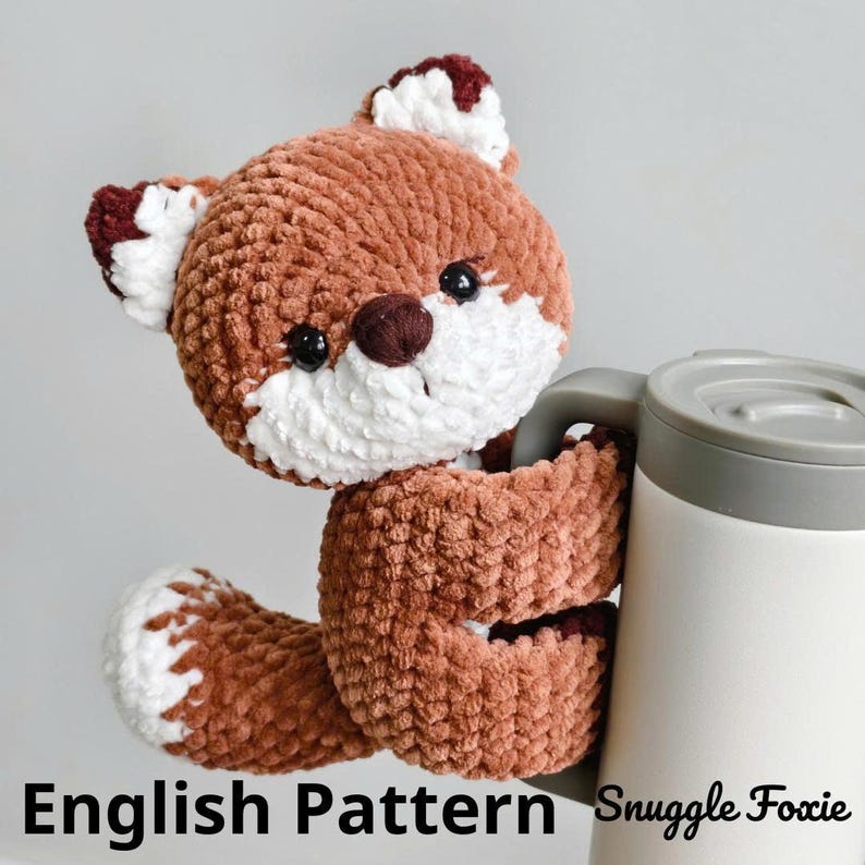 May include: A crocheted fox plushie in shades of brown, white, and burgundy, holding a white and gray travel mug. The fox has black eyes and a brown nose. The text "English Pattern Snuggle Foxie" is at the bottom.