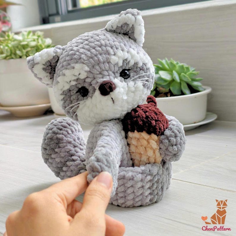K&ouml;nnte beinhalten: Ein geh&auml;keltes Eichh&ouml;rnchen-Pl&uuml;schtier in Grau und Wei&szlig;, das eine braune und beige Eichel h&auml;lt. Das Eichh&ouml;rnchen hat schwarze Augen und eine braune Nase. Das Pl&uuml;schtier ist aus weichem, strukturiertem Garn gefertigt.