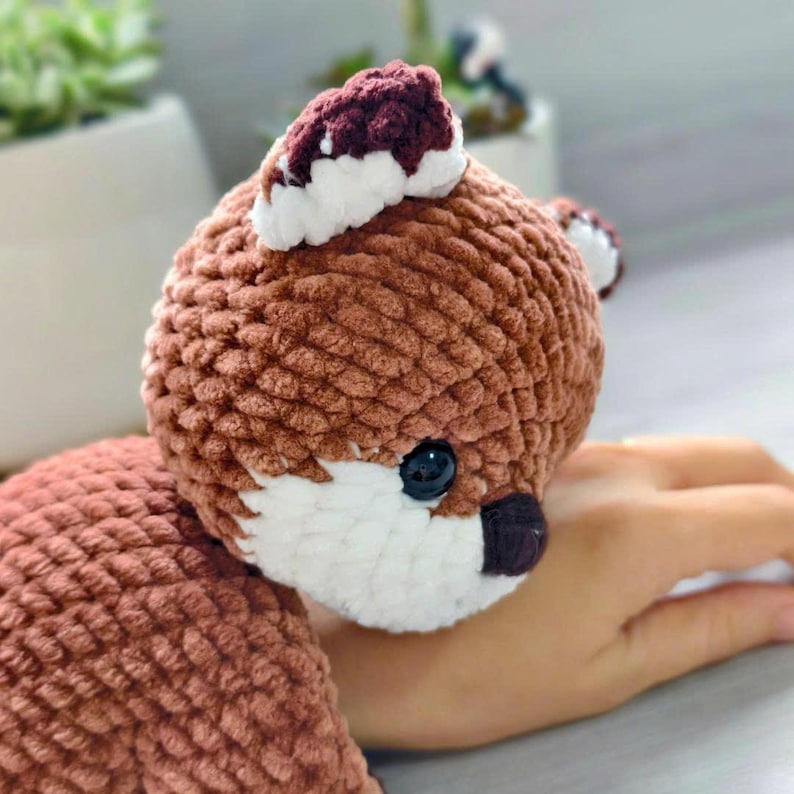 May include: A handmade, plush fox toy in shades of brown, white, and burgundy. The fox has a round head with a white face, black eyes, and a burgundy nose. The ears are burgundy with white tips. The toy is soft and cuddly.