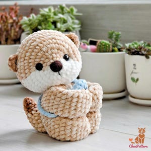 May include: A crocheted otter plush toy in tan, brown, and white. The otter has black eyes, a brown nose, and a blue accent. The plushie is sitting with its arms wrapped around itself. The background includes potted plants.