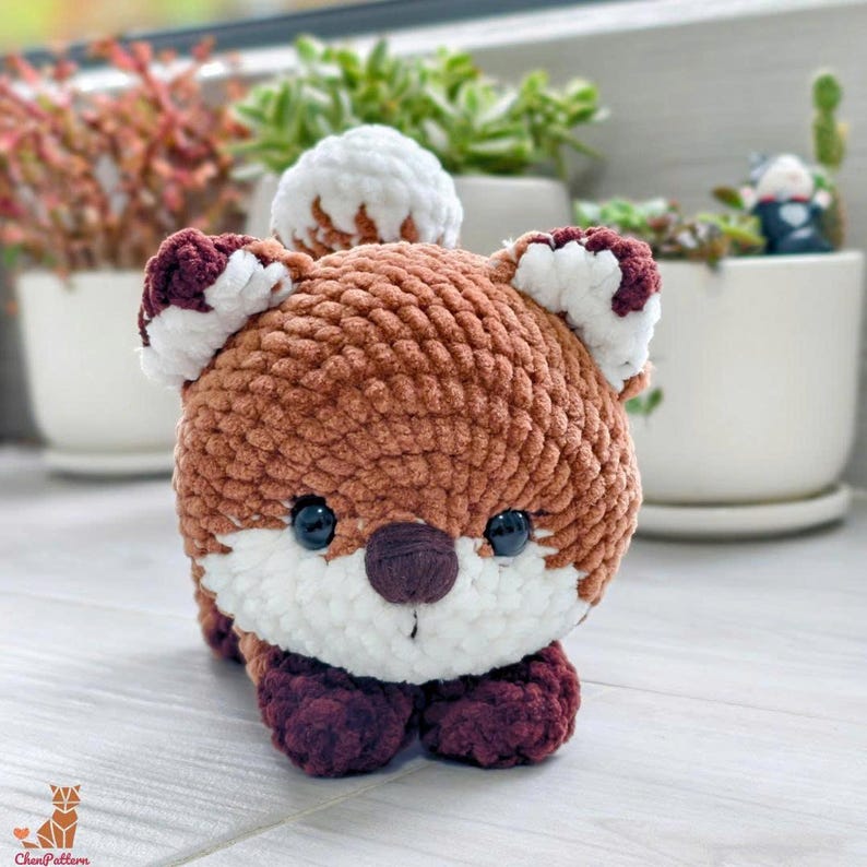 May include: A handmade, crocheted fox plushie in shades of brown, white, and burgundy. The plush toy has large black eyes, a white muzzle, and burgundy paws and ear accents. The fox is lying down, showcasing its soft, textured design.