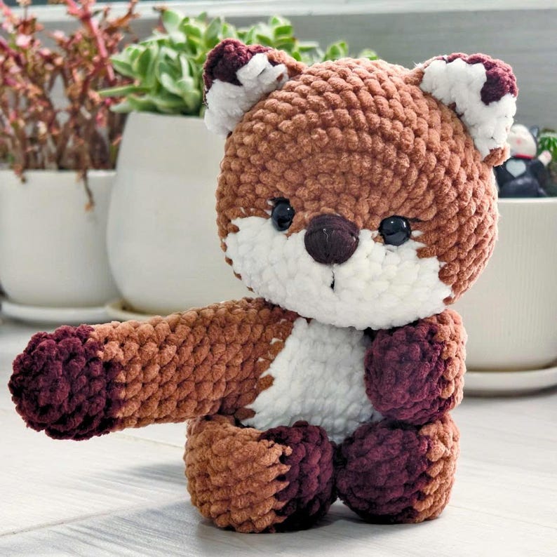 May include: A handmade crochet fox toy in shades of brown, white, and burgundy. The plush fox has a rounded body, a white chest, and a long arm extended to the left. The fox has black eyes and a dark brown nose.