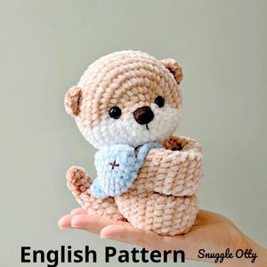May include: A handmade, plush otter toy in shades of tan, white, and brown. The otter has a light blue bib with a cross design. The text "English Pattern" is visible at the bottom. The toy is made of soft, textured material.