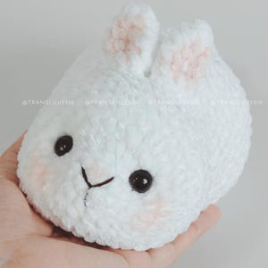 May include: A white plush toy shaped like a round bunny with soft, textured yarn. It features two upright ears with light pink centers, two black plastic eyes, a stitched black nose and mouth, and faint pink blush marks on its cheeks. Text reads @TRANGLUU2310.