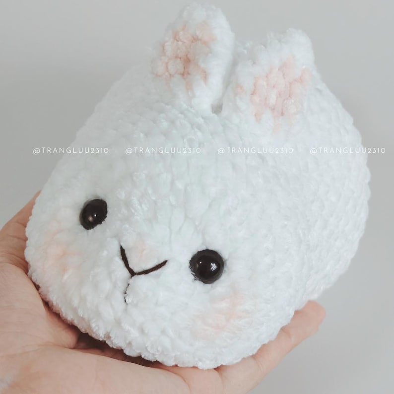 May include: A white plush toy shaped like a round bunny with soft, textured yarn. It features two upright ears with light pink centers, two black plastic eyes, a stitched black nose and mouth, and faint pink blush marks on its cheeks. Text reads @TRANGLUU2310.
