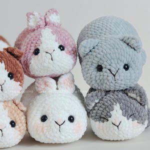 May include: A collection of five plush crochet animals made from soft, fuzzy yarn. The group includes a pink and white rabbit, a solid white rabbit, a light gray cat, a dark gray and white cat, and a brown and white animal, all with black eyes.
