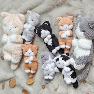 Op de afbeelding: Een verzameling van zeven gehaakte pluche kattenspeeltjes in diverse kleuren, waaronder crème, lichtgrijs, zwart-wit en oranje. Elk knuffeldier heeft een slapende uitdrukking met gesloten ogen, kleine oortjes en witte pootjes van zacht garen.