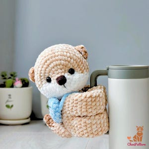 May include: A crocheted teddy bear in beige and white, with black eyes and a brown nose, wearing a light blue scarf. The bear is holding a white travel mug with a grey handle. The mug has a logo on the bottom right. The mug is likely to hold 400 ml.