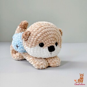 May include: A crocheted otter toy in tan, white, and brown, with black eyes and a black nose. The otter is wearing a light blue nappy with a black cross. The toy is lying down.
