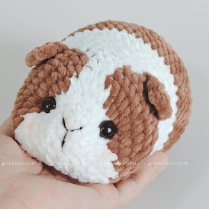 May include: A small, round, crocheted plush toy shaped like a guinea pig. The plush features soft, textured yarn in alternating patches of brown and white. It has two shiny black plastic eyes and a stitched nose detail on the white face area.