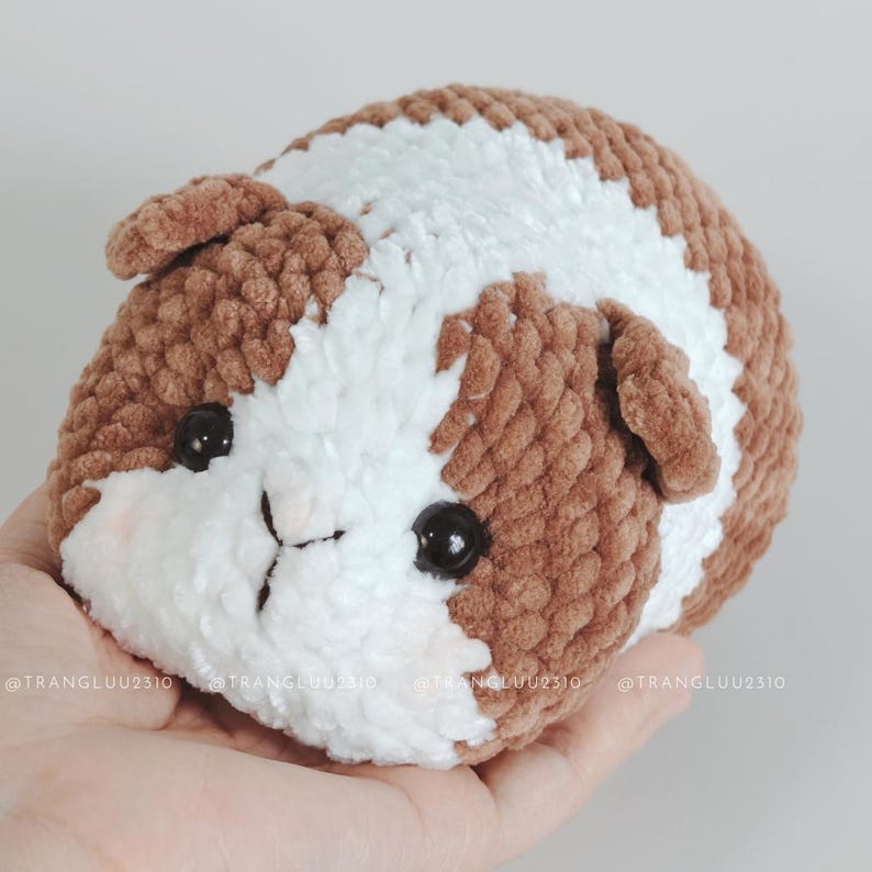 May include: A small, round, crocheted plush toy shaped like a guinea pig. The plush features soft, textured yarn in alternating patches of brown and white. It has two shiny black plastic eyes and a stitched nose detail on the white face area.