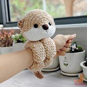 May include: A handmade plush otter toy in shades of beige and white. The otter has black eyes and a brown nose. The plush toy is designed to wrap around a wrist. The toy is made of a soft, textured material.
