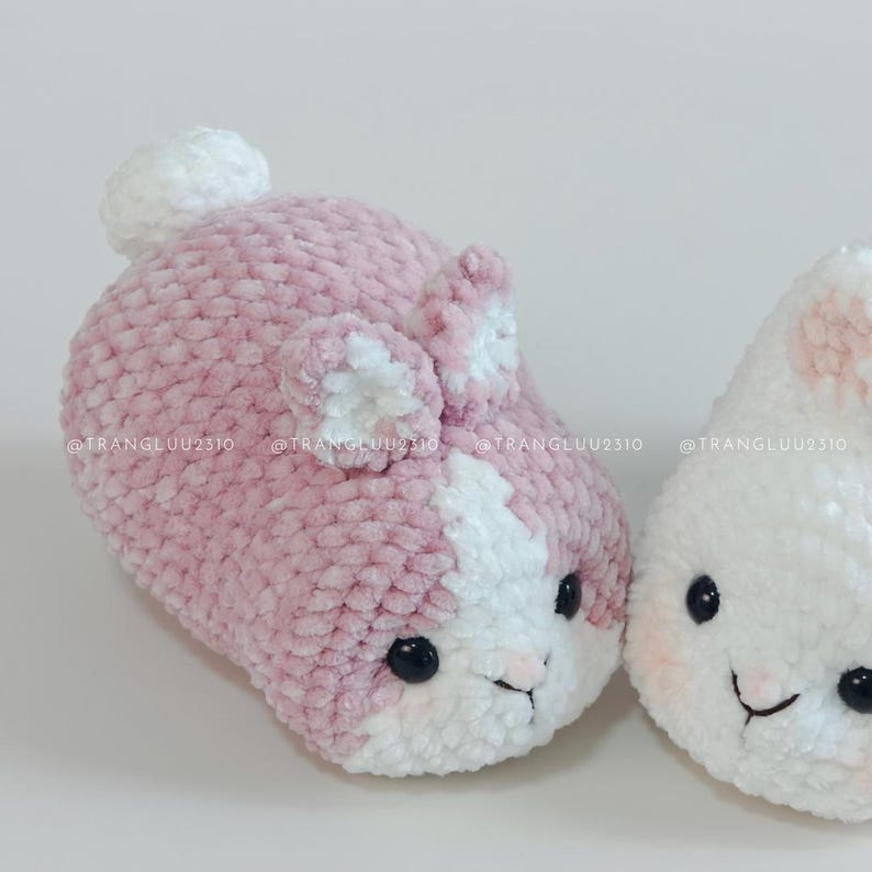 May include: Two plush crochet animals made of soft, dusty pink and white yarn. The left animal features a white tail, two upright ears, and black bead eyes. The right animal is primarily white with pink accents. Text reads @TRANGLUU2310.
