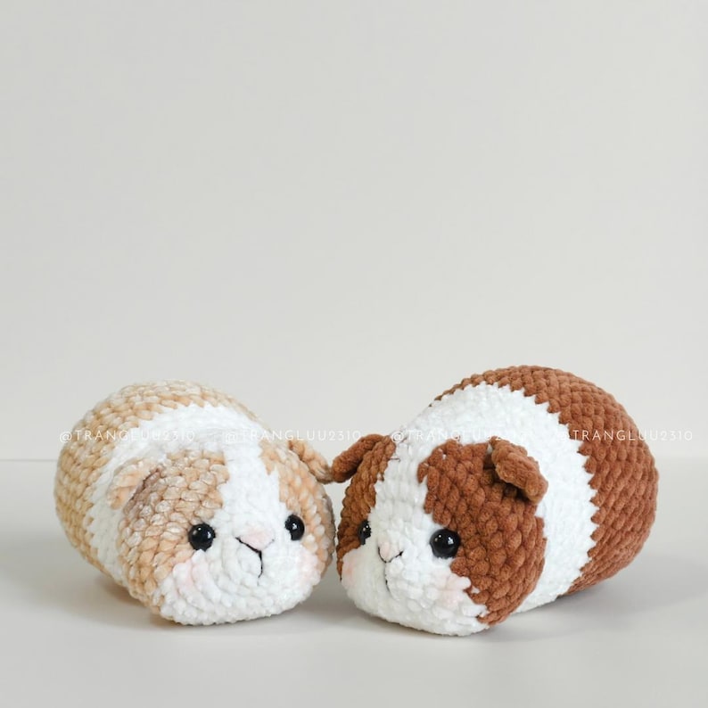 May include: Two plush guinea pig toys made of soft yarn. One toy features light tan and white fur, while the other has dark brown and white fur. Both have small black eyes, stitched noses, and subtle pink blush details on their cheeks.