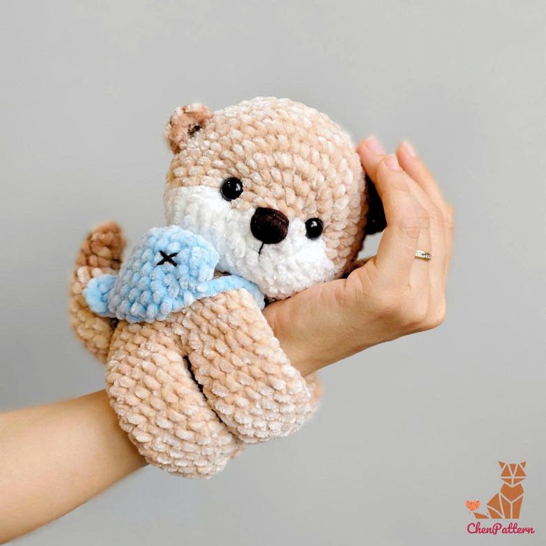 May include: A handmade plush otter toy in shades of tan, white, and brown, with a light blue scarf. The otter has black eyes and a brown nose. The toy is held in a hand against a grey background.