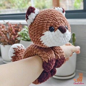 May include: A handmade crochet fox toy in shades of brown, white, and burgundy. The fox has a round head with black eyes, white accents, and a fluffy tail. The toy is designed to be worn on the wrist.