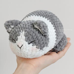 May include: A small, plush, crocheted cat toy featuring a soft, textured yarn in shades of grey and white. The cat has a rounded body, small ears, and closed eyes with a stitched nose. The text @TRANGLUU2310 is visible across the surface.