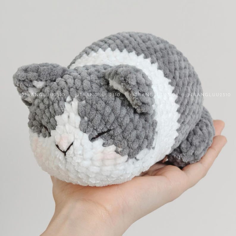 May include: A small, plush, crocheted cat toy featuring a soft, textured yarn in shades of grey and white. The cat has a rounded body, small ears, and closed eyes with a stitched nose. The text @TRANGLUU2310 is visible across the surface.