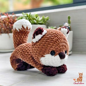 May include: A handmade crochet fox toy in shades of brown, white, and burgundy. The plush fox has a fluffy tail, white accents on its ears and face, and dark eyes. The fox is lying down, showcasing its detailed craftsmanship.