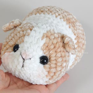 May include: A plush guinea pig toy crafted from soft, textured yarn in shades of beige and white. The stuffed animal features two round, glossy black eyes, a small pink stitched nose, and two small ears on the sides of its rounded, compact body.