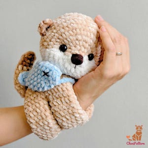 May include: A handmade, plush otter toy in shades of tan, white, and brown, with a light blue accent. The otter has black eyes and a dark brown nose. The toy is held in a hand, showcasing its size and texture. The toy is a cute, cuddly item.
