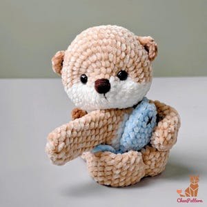 May include: A handmade, plush otter toy in shades of tan, brown, and white. The otter has black eyes, a brown nose, and a light blue fish. The toy is made of soft, textured yarn and is sitting down.
