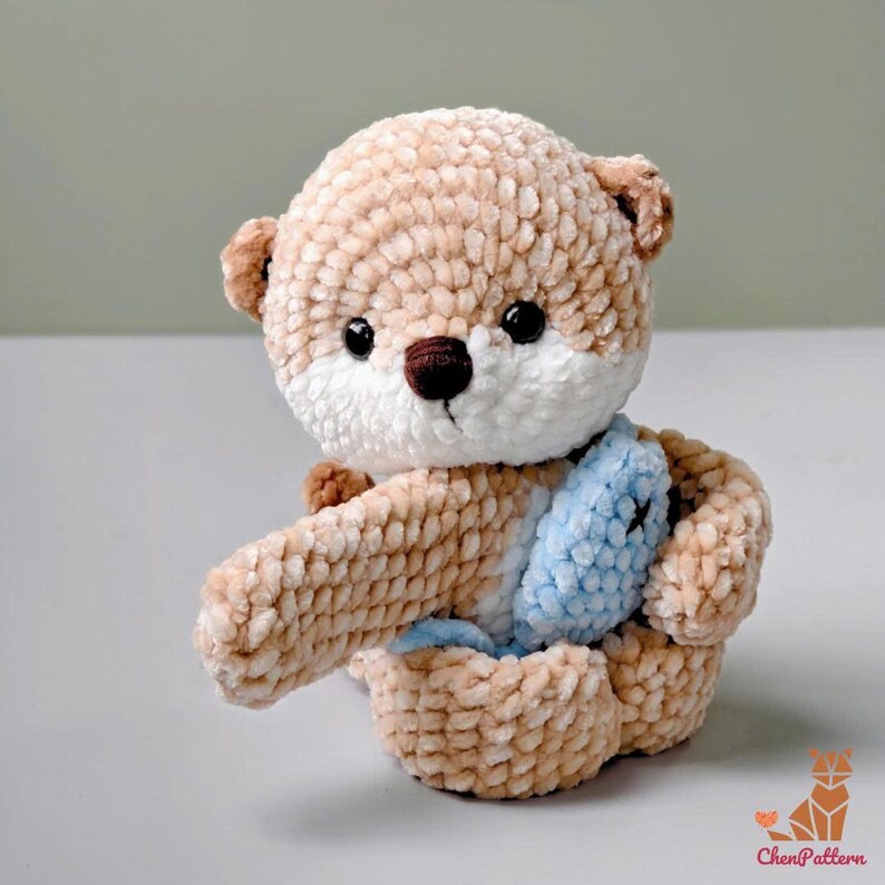 May include: A handmade, plush otter toy in shades of tan, brown, and white. The otter has black eyes, a brown nose, and a light blue fish. The toy is made of soft, textured yarn and is sitting down.