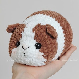 May include: A plush guinea pig toy made of soft brown and white yarn with a textured, fuzzy appearance. The toy features two small black plastic eyes, a stitched nose, and subtle pink blush details on the cheeks. The text @TRANGLUU2310 is visible.