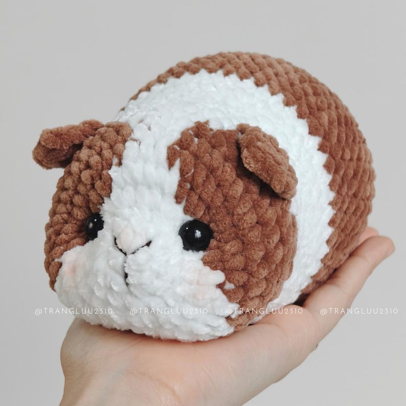 May include: A plush guinea pig toy made of soft brown and white yarn with a textured, fuzzy appearance. The toy features two small black plastic eyes, a stitched nose, and subtle pink blush details on the cheeks. The text @TRANGLUU2310 is visible.