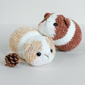 May include: Two round, plush crochet guinea pig toys with soft yarn texture. One is white and light tan, the other is white and brown. Both have black plastic eyes, small ears, and pink blush details. A small brown pinecone sits beside them. @TRANGLUU2310