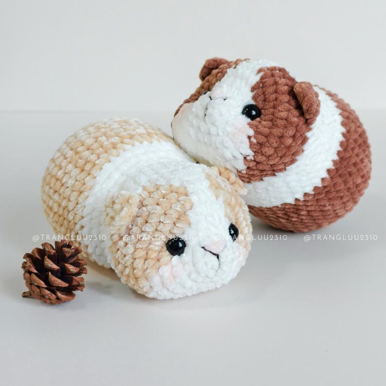May include: Two round, plush crochet guinea pig toys with soft yarn texture. One is white and light tan, the other is white and brown. Both have black plastic eyes, small ears, and pink blush details. A small brown pinecone sits beside them. @TRANGLUU2310