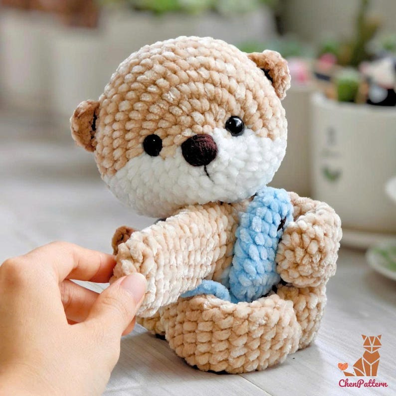 May include: A crocheted teddy bear in tan and white, with black eyes and a brown nose. The bear sits in a matching crochet basket, with a blue accent. The bear's ears and paws are a darker shade of brown. The bear is handmade.