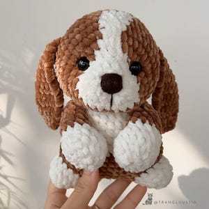 May include: A handmade plush puppy with brown and white fur. The dog has black eyes and a brown nose. The plush toy is made of soft, textured yarn. The ears and body are brown, with white accents on the face and paws. A cute, cuddly stuffed animal.