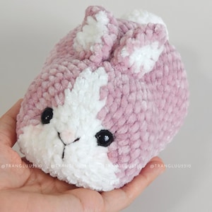 May include: A small, round, plush crochet bunny made of soft, dusty pink and white yarn. The bunny features two upright ears, a white patch on its face, and two shiny black plastic eyes. The texture is fuzzy and thick, creating a rounded shape.