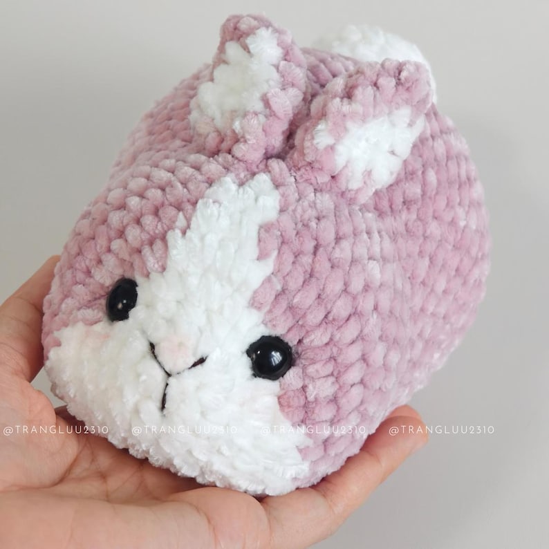 May include: A small, round, plush crochet bunny made of soft, dusty pink and white yarn. The bunny features two upright ears, a white patch on its face, and two shiny black plastic eyes. The texture is fuzzy and thick, creating a rounded shape.