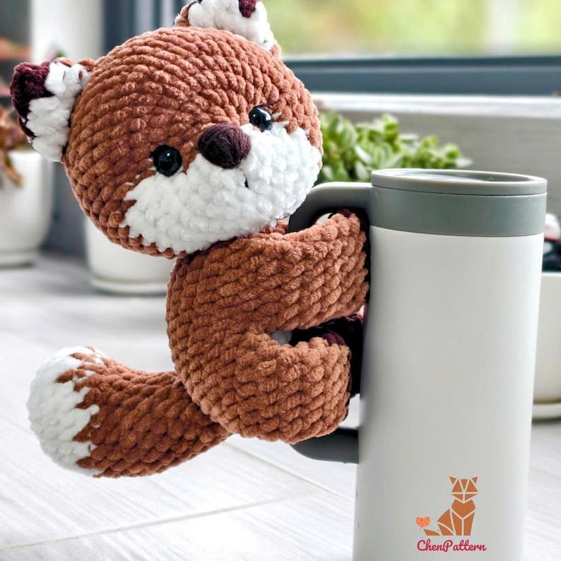 May include: A handmade, brown and white crocheted fox toy hugging a white travel mug with a gray handle. The fox has black eyes and a white muzzle. The mug has a green lid and a fox logo with the text "ChenPattern".